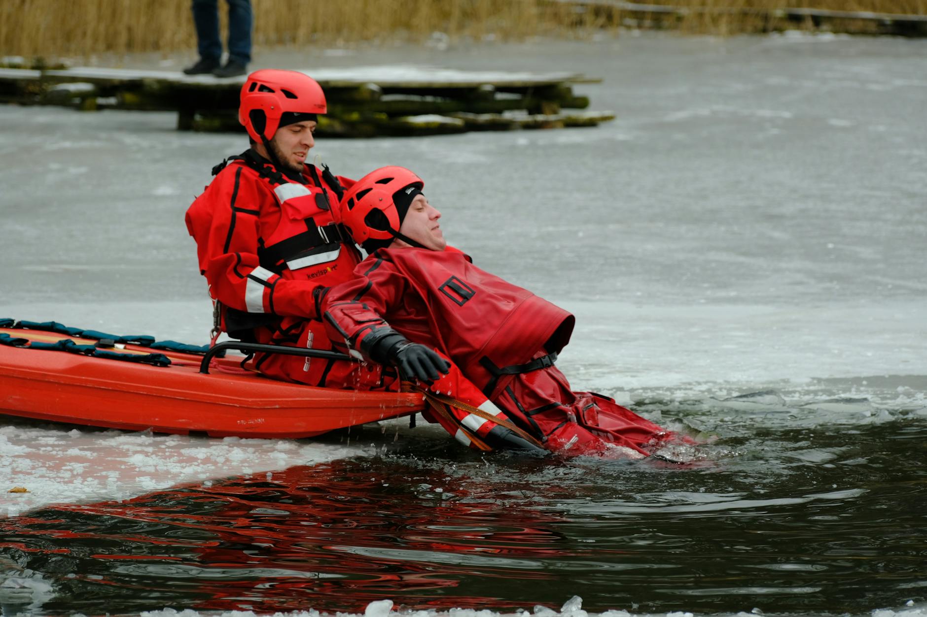 Dwóch ratowników w czerwonych kombinezonach ćwiczy akcję na zamarzniętym jeziorz